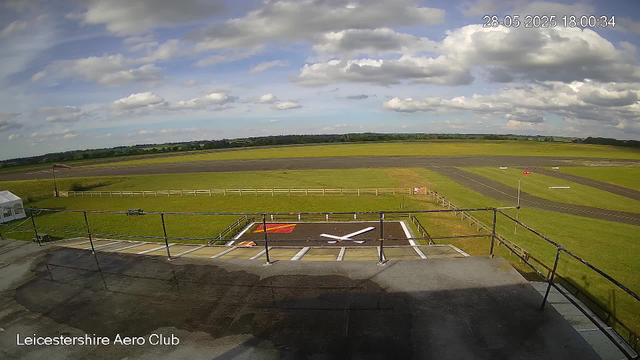 A view from a webcam at Leicestershire Aero Club shows an airfield with a runway and grassy areas. The sky is partly cloudy with patches of blue. In the foreground, there is a helipad marked with a white "H" and a red and yellow flag beside it. A small white tent is visible on the left side, and the airfield is enclosed by a white fence. The timestamp in the upper right corner indicates the date and time as May 28, 2025, at 6:00 PM.