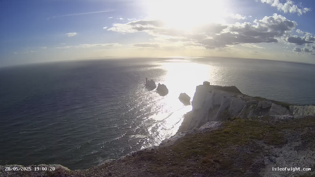 A view of a rugged coastline with white chalk cliffs and three sea stacks extending into the ocean. The sun is shining brightly, reflecting off the water, and the sky is partly cloudy. The landscape appears serene with gentle waves washing against the shore, and the time stamp indicates the photo was taken on May 28, 2025, at 19:00.