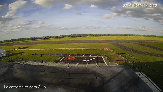 This image shows a view of an airfield with a large clock painted on the ground in the foreground. The clock has white hands and a red square background. Beyond the fenced area, the runway extends into the distance, surrounded by green grass and open fields under a partly cloudy sky. The date and time in the top right corner indicate 28th May 2025, 7:00 PM. The bottom left corner shows the text "Leicestershire Aero Club."