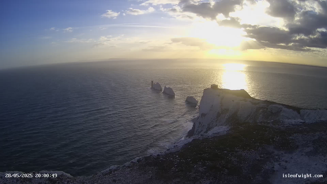 A view of white chalk cliffs and rock formations jutting out from the ocean at sunset, with the sun low in the sky casting a golden glow over the water and clouds. The date and time are displayed in the bottom left corner, and a website URL is in the bottom right corner.