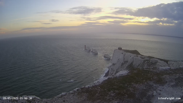 A panoramic view of white chalk cliffs rising sharply from the ocean, with some of the cliffs topped with patches of grass. There are large, broken sections of chalk falling into the water below. The sky is partly cloudy with the sun setting or rising on the horizon, casting a soft, warm light over the scene. A small structure is visible on the highest point of the cliff on the right. The ocean appears calm, with gentle waves. The timestamp in the lower left corner reads "28/05/2025 21:00:30" and the website "isleofwight.com" is displayed in the lower right corner.