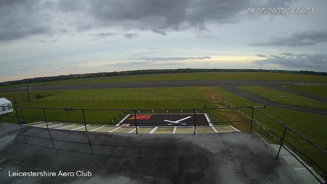 A view from a webcam at Leicestershire Aero Club shows an overcast sky with dark clouds. In the foreground, there is a helicopter landing pad marked with a large white "H" on a black background, surrounded by a fence. The tarmac appears wet. To the left, part of a small white tent and some equipment are visible. In the background, there are green fields and a paved runway stretching horizontally across the image. The timestamp in the top right corner reads "28-05-2025 21:00:51".