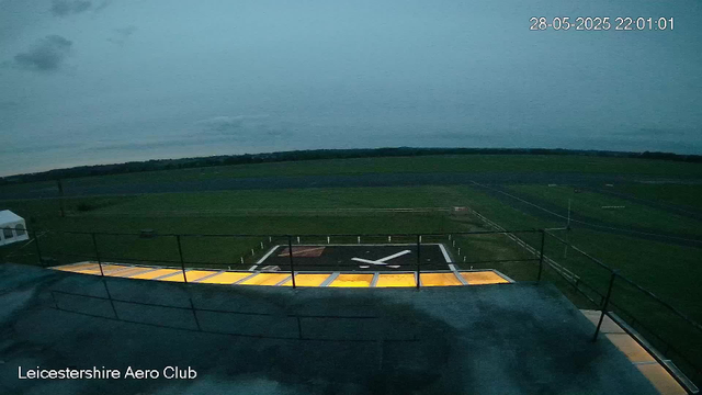 An outdoor view from a webcam shows a green open field with a runway or helipad marked with a large black "X" and yellow-lit panels along its edges. A black railing surrounds the viewing area. The sky is mostly cloudy with a few patches of light, and the timestamp indicates it is early evening on May 28, 2025. The words "Leicestershire Aero Club" appear at the bottom of the image.