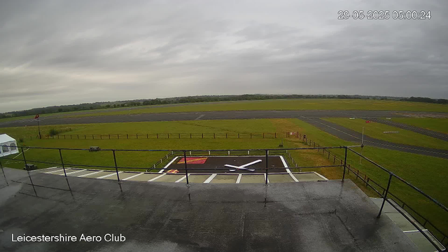 A wide view from a webcam shows the runway and surrounding grassy area at Leicestershire Aero Club. The sky is cloudy and overcast. In the foreground, there's a large helicopter landing pad with painted markings, including a red and yellow symbol. The runway extends into the distance, with a few signs and navigational aids visible. The scene appears to be taken early in the morning, as indicated by the timestamp.