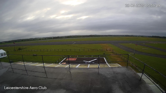 A view from a webcam at Leicesterhire Aero Club showing a cloudy sky over an empty runway and grassy areas. In the foreground, there is a clock painted on a small section of the pavement, with the hands pointing to approximately 10:15. A railing is visible at the edge of the platform, and a white tent is on the left side. The scene appears to be early morning with overcast weather. The timestamp in the upper right corner reads 29-05-2025 05:59:49.