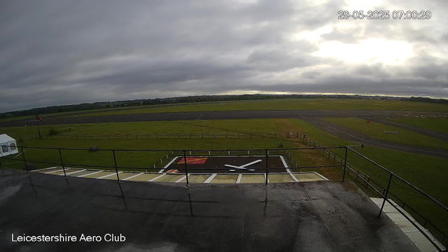 A view of an airport runway and tarmac area on a cloudy day, with a large clock painted on the ground and a windsock visible on the left side. The timestamp on the image reads "29-05-2025 07:00:29," and the location is indicated as "Leicestershire Aero Club." The scene shows an open, grassy landscape stretching out into the distance under a mostly cloudy sky.
