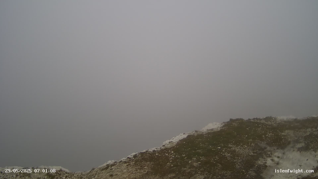 A foggy, overcast sky with thick gray clouds dominates the scene. The lower part of the image shows a rocky, grassy hill with some patches of white, possibly snow or frost, and an area that appears wet or muddy. There are no visible trees or structures, and the overall atmosphere looks cold and misty. The timestamp in the bottom left corner reads "29/05/2025 07:01:08," and the bottom right corner has the website "isleofwight.com."