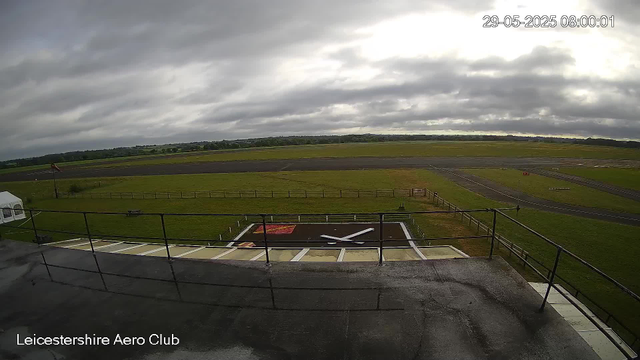 A view from a webcam at Leicestershire Aero Club shows a large open airfield with a runway extending across the landscape. In the foreground, there is a helipad with a white "H" painted on it, surrounded by a fence, and a small structure on the left side. The sky is overcast with dark, heavy clouds, and the landscape features grassy areas and distant trees on the horizon. The timestamp in the top right corner indicates the date and time as May 29, 2025, at 8:00 AM.