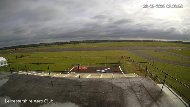A view from a webcam showing the Leicester Aero Club with a windsock, a small helipad with a white "H," and a grassy airfield area. The sky is cloudy with patches of sunlight, and distant trees and open fields extend towards the horizon. The timestamp indicates the date and time as May 29, 2025, at 09:00:56.