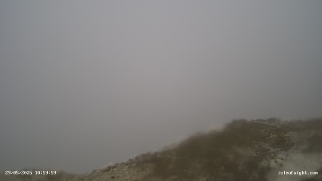 A landscape view from a webcam showing a foggy or overcast sky with limited visibility. The ground appears rocky and barren at the bottom right corner. The timestamp on the image indicates it was taken on May 29, 2025, at 10:59:59. The website "isleofwight.com" is also visible in the bottom right corner.