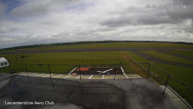 An outdoor view from a webcam at Leicesterhire Aero Club showing an overcast sky with clouds. In the foreground, there is a large clock monument lying flat on the ground, with the time showing approximately 10:10. A small flagpole with a British flag is visible on the left side. The scene includes a fenced grassy area and a runway extending into the distance, with some runway markings and small structures. The image is timestamped 29-05-2025 11:00:54.