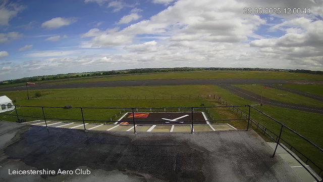 A wide view of an airfield under a partly cloudy sky. In the foreground, there's a rooftop with a railing, a helipad with a white "H" marked on it, and a small tent. Beyond the helipad, there's a grassy area with a flagpole flying a small flag. The airfield has runways, taxiways, and some equipment spread across the tarmac. The horizon shows a line of trees and distant fields. The image is timestamped "29-05-2025 12:00:41" and labeled "Leicestershire Aero Club."