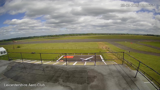 An outdoor view of an airstrip at Leicestershire Aero Club, with a small helipad marked on the ground. The sky is partly cloudy with patches of blue, and the landscape extends into open fields with distant trees on the horizon. There is a windsock on the left side of the image, and the foreground shows a parking area with a metal railing. The timestamp in the upper right corner indicates the date as May 29, 2025, at 12:59:57.