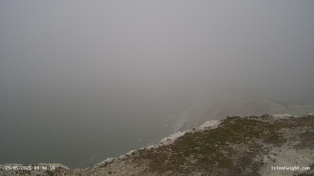 A foggy, mountainous landscape with limited visibility. The foreground shows a rocky and grassy edge, while the background is obscured by thick fog, blending the sky and landscape into a uniform grayish mist.