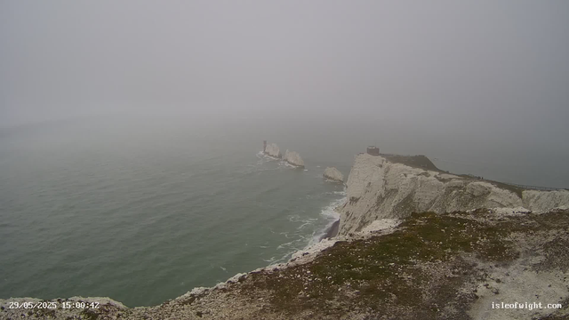 A foggy view of white chalk cliffs along a coastline with a small building on top of one of the cliffs. The ocean is visible below with gentle waves, and there's a footpath along the edge of the cliffs. A lighthouse or lighthouse-like structure is at a distance on a rock formation in the water. The image has a timestamp of "29/05/2025 15:00:42" and a website URL "isleofwight.com" at the bottom right corner.