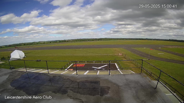 A view from a webcam showing an overcast sky with scattered clouds over a flat airfield. In the foreground, there is a helipad with a large white "H" painted on it, surrounded by a metal railing and some markings. To the left of the helipad, a small tent or canopy is visible, covered with a protective white tarp. The airfield has a paved taxiway and grassy areas, with a distant horizon of trees and fields. The image is timestamped 29-05-2025 at 15:00:41, and the location is indicated as Leicestershire Aero Club.