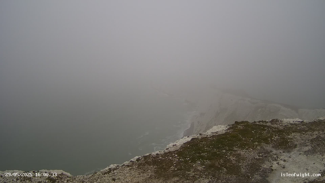 A foggy scene over a coastal cliff with the ocean visible below. The visibility is low due to dense fog, obscuring much of the background and horizon. The foreground shows a rocky, uneven surface with patches of moss or grass. The date and time stamp in the lower left corner reads "29/05/2025 16:00:33," and the bottom right corner has the website "isleofwight.com."
