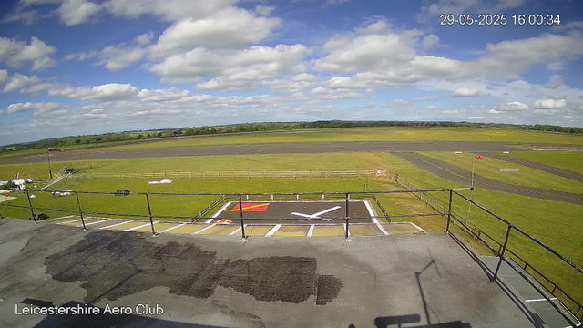 A view from a webcam at Leicestershire Aero Club shows a paved area with a large helicopter landing pad marked with a white "H" on a red background and surrounding white lines. In the background, there is a grassy field enclosed by a wooden fence, and beyond that, an open expanse of fields and sky. The sky is partly cloudy with patches of blue. The timestamp indicates the date and time as May 29, 2025, at 4:00 PM and 34 seconds.