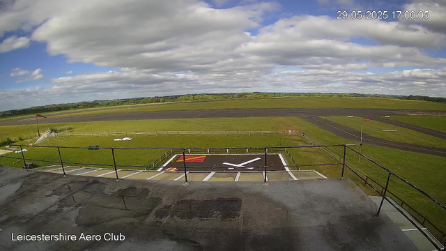 A wide view of an outdoor airfield at Leicester Aeroclub. In the foreground, there is a helicopter landing pad marked with a large white "X" and surrounded by a black railing. To the left, a flagpole with a red flag, and some small structures and equipment near a white railing. Beyond the pavement, there is green grass and a running track with white lines extending into the distance. The sky is partly cloudy with patches of blue, and the date and time are displayed in the upper right corner: 29-05-2025 17:00:05. The bottom left corner has text reading "Leicestershire Aero Club."