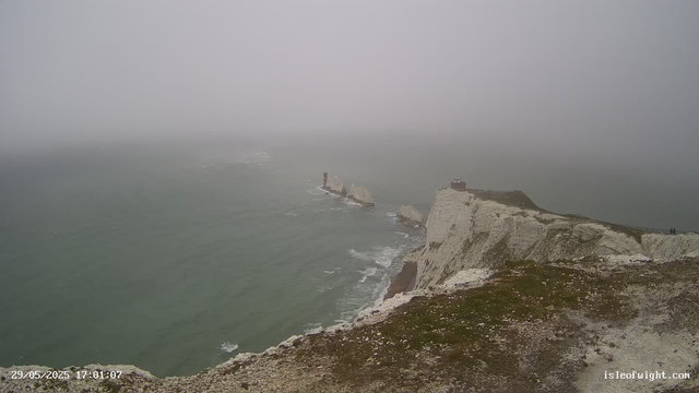 A foggy view of white chalk cliffs along the coastline with a narrow footpath at the top. The ocean waves crash against the base of the cliffs, and distant rock formations and a lighthouse are visible in the fog. The scene appears to be taken during late afternoon or early evening.