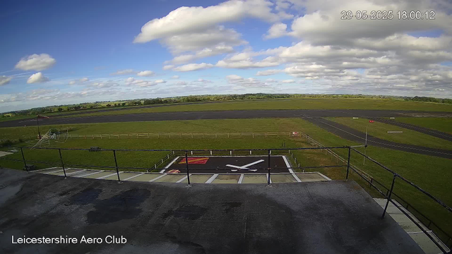 A wide view of an airfield shows a runway in the background under a partly cloudy sky. In the foreground, there is a helipad marked with a large white "H" and a red square, surrounded by a black asphalt surface. A metal railing runs along the edge of a platform or balcony, and a small flag is visible on the left side. The landscape beyond the airfield is flat and green with fields and trees in the distance. The image includes a timestamp in the upper right corner (29-05-2025 18:00:12) and the text "Leicestershire Aero Club" at the bottom left corner.