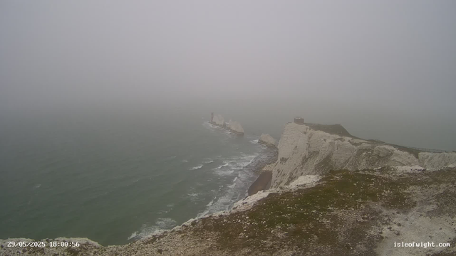 A foggy view of white cliffs along a rocky coastline with two prominent sea stacks in the distance, partly obscured by mist. The sea below appears choppy, and the sky is overcast, creating a moody atmosphere. A small building or structure is visible on the top of the rightmost cliff. The timestamp indicates it was taken on May 29, 2025, at 6:00 PM.