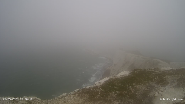 A foggy coastal scene with cliffs and the ocean below. Visibility is low due to dense fog, obscuring much of the landscape. The rugged cliffs descend into the sea, with waves crashing against the rocks. The image is timestamped "29/05/2025 19:00:44" and is from isleofwight.com.