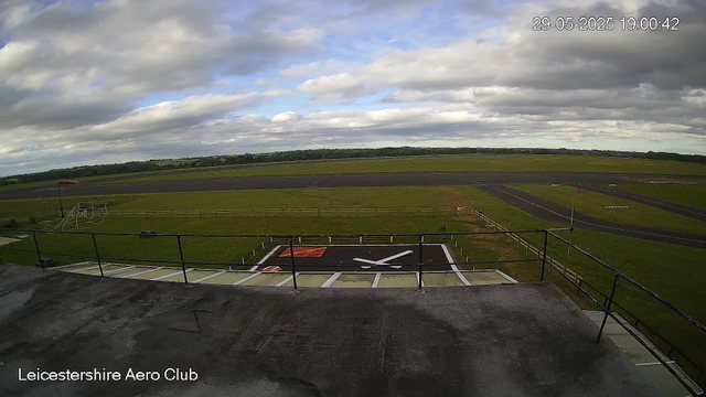 An outdoor view from a webcam at Leicesterhire Aero Club shows the airfield under a partly cloudy sky. In the foreground, there is a rooftop with some markings and a railing. A windsock is visible near the center-left, along with a small fenced area. The runways and taxiways extend into the distance, surrounded by green grass. The horizon features a line of trees, and the date and time are displayed at the top right corner: 29-05-2025, 19:00:42.