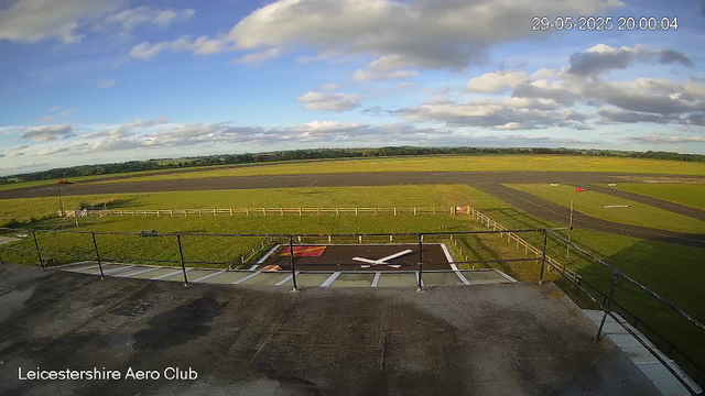 A wide view of Leicester Aero Club’s airfield under a partly cloudy sky, with green fields, a runway in the distance, and a helipad with a large white "X" on it in the foreground. The photo is timestamped "29-05-2025 20:00:04" and shows a railing along the edge of a rooftop or observation platform.