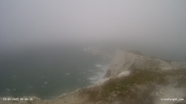 A foggy view of the coastline with white chalk cliffs and the ocean waves crashing against the rocks, partially obscured by thick fog. A lighthouse is barely visible in the distance. The timestamp reads "29/05/2025 20:00:34" and the website "isleofwight.com" is displayed in the bottom right corner.