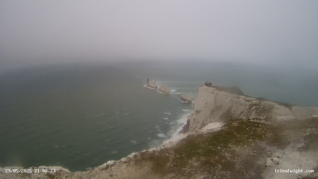 A view of the white chalk cliffs along the coastline, with a lighthouse on top of one of the cliffs. The sea below appears rough with waves, and the sky is overcast and foggy, creating a moody atmosphere.