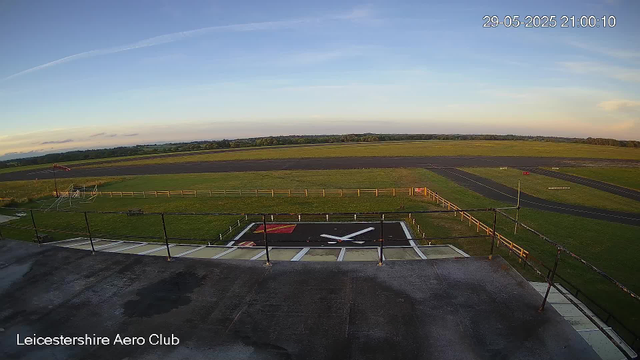 An outdoor view from a webcam at Leicestershire Aero Club showing a large clock on the ground, a grassy field with a small flagpole and windsock on the left, a runway stretching across the middle distance, and a wide open sky with some clouds and a contrail. The timestamp on the image is 29-05-2025 21:00:10.