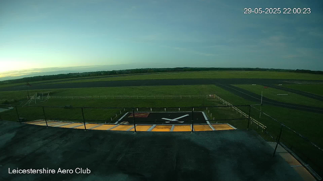 An outdoor view from a webcam at Leicestershire Aero Club showing a helicopter landing pad with a large black and yellow area, a white 'X' marking, and surrounding black railings. In the background, there are green fields, a few small structures, a paved runway, and a clear blue sky with a hint of sunrise or sunset on the horizon. The timestamp shows the date and time as 29-05-2025 22:00:23.
