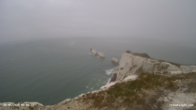 A view of a foggy seacoast with white chalk cliffs and rocky outcrops extending into the sea. The ocean appears calm with gentle waves, and the sky is overcast, creating a muted, gray atmosphere. A small lighthouse or navigation marker is visible on top of a distant rocky island. The timestamp shows "30/05/2025 05:00:15" and the website "isleofwight.com" is displayed in the lower right corner.