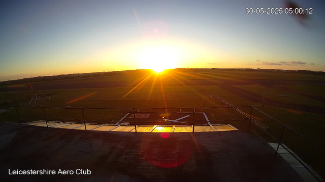 A sunrise over an airfield, with the sun partially above the horizon casting a golden glow across the tarmac and surrounding grassy fields. The sky is mostly clear with a few scattered clouds. Visible are a helipad, some runway markings, and fencing along the edge of the airfield. The timestamp indicates the image was taken early in the morning.