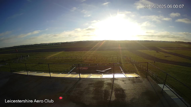 The image shows a bright, sunny morning at Leicester Aeroclub with the sun low in the sky, partly cloudy, and fields stretching into the distance. The foreground includes a small platform or area with railings, and a windsock is visible on the left side. The scene is peaceful with open grassy fields and a distant horizon. There is a timestamp in the top right corner indicating the date and time as May 30, 2025, at 6:00 AM. The bottom left corner displays the text "Leicestershire Aero Club."