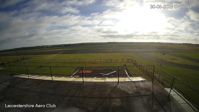An outdoor scene at the Leicestershire Aero Club on a partly cloudy day. The image shows a grassy airfield with runways in the distance, a small helipad or tarmac area with some equipment, and a fence surrounding the area. The sky is partly cloudy with the sun shining through the clouds. The digital timestamp indicates the date and time as 30th May 2025 at 07:00:01.