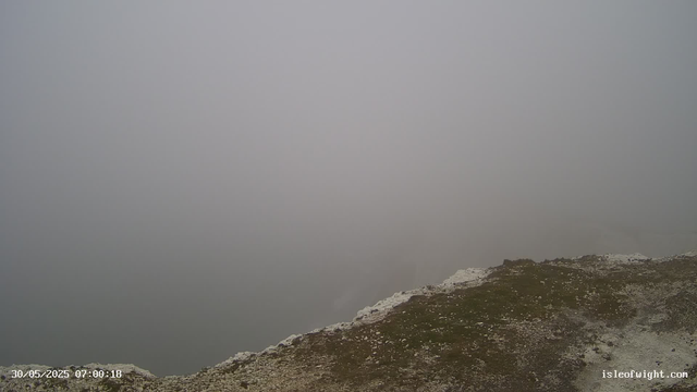 A foggy mountain landscape with a rocky and grassy ridge at the bottom. The sky is overcast with thick fog obscuring the view beyond the ridge. There is a timestamp in the lower left corner indicating the date and time as 30/05/2025 07:00:18, and a website URL "isleofwight.com" in the lower right corner.