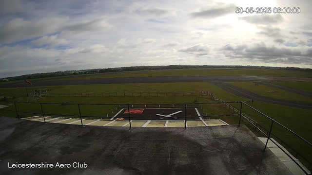 An outdoor view from a webcam at Leicesterhire Aero Club shows a cloudy sky overhead. In the foreground, there is a helipad with markings and a black railing around it. To the left, a small flagpole with a flag and a basketball hoop are visible. Beyond, there is a large open grassy area with a fence and a runway extending into the distance. The timestamp in the top right corner indicates the date and time as 30-05-2025 08:00:00.