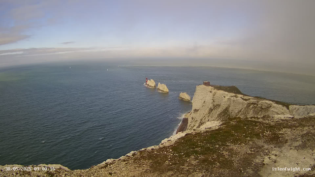 A view from a webcam showing white chalk cliffs along the coast extending into the distance over the sea. There are several large rock formations just offshore, with one featuring a small red and white lighthouse. The sky is partly cloudy with patches of blue, and the water appears calm. The timestamp indicates the photo was taken on May 30, 2025, at 9:00 AM.