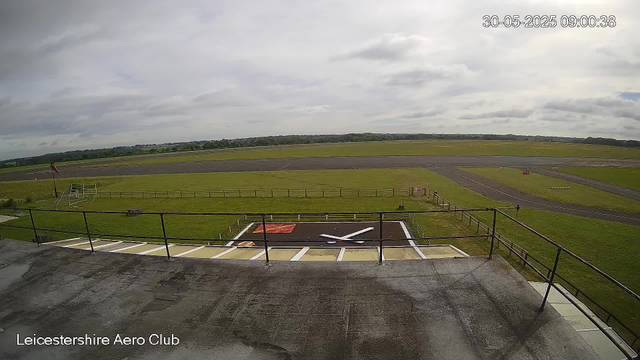 An outdoor view from a webcam at Leicestershire Aero Club shows an empty grassy airfield with a small helicopter landing pad and a windsock on the left. In the background, there are runways and a cloudy sky, with rolling green fields and distant trees beyond the airfield. The timestamp indicates the photo was taken on May 30, 2025, at 9:00 AM.