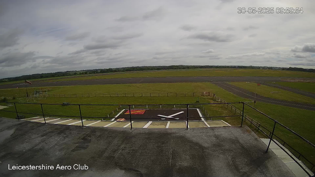 An overcast sky above an empty runway on a cloudy day at Leicester Aero Club. The camera captures a helipad with a large white "X" and orange markings in the foreground, a grassy area with a windsock to the left, and the main runway stretching across the middle distance. Rolling green fields extend into the horizon under a cloudy sky.