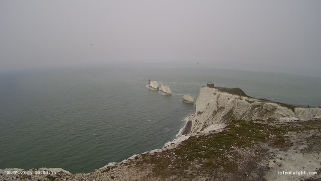 Overcast sky with foggy conditions over a calm ocean. The white chalk cliffs extend into the sea, with a small building and a red and white lighthouse visible on the edge of the cliffs. A narrow pathway runs along the top of the cliffs with a few small figures walking. The date and time are displayed at the bottom left corner, and a website URL is at the bottom right corner.