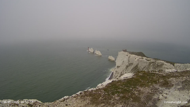 A view of white chalk cliffs extending into the ocean under a foggy sky. The cliffs have some grassy patches on top, and there are small buildings or structures near the edge. The water below is calm, with gentle ripples. The scene is foggy, obscuring the horizon and creating a muted, misty atmosphere. The timestamp on the lower left shows "30/05/2025 10:59:59," and the website "isleofwight.com" is visible in the lower right corner.