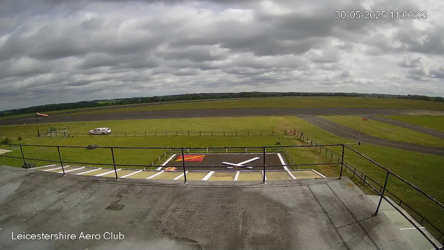 A view from a webcam at Leicestershire Aero Club shows a cloudy sky over an empty airfield with a runway in the distance. In the foreground, there is a helipad with painted markings, a safety railing, and part of the building’s roof. A white vehicle is parked on a grassy area near a flagpole with a red and white flag. The surrounding landscape is open with green fields and distant trees under a sky filled with dark, heavy clouds. The timestamp reads "30-05-2025 11:00:03."