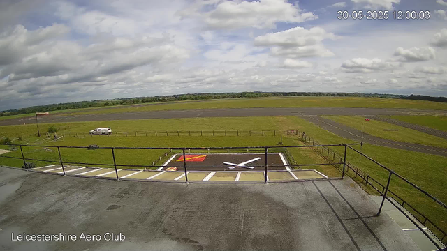 A scenic view from a webcam at Leicestershire Aero Club shows a cloudy sky over an open grassy area with a small runway in the background. In the foreground, there is a parking area with white lines and a helipad marked with a large white "H" on a dark surface. A white vehicle is parked on the grass near a flag. The horizon is lined with trees and fields, and the date and time displayed in the top right corner is 30-05-2025 at 12:00:03. The bottom left corner of the image contains the text "Leicestershire Aero Club."