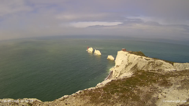 A view of layered white chalk cliffs extending into the sea with a lighthouse on one of the promontories, under a sky with scattered clouds. The ocean has gentle waves, and the coastline curves as the cliffs stretch into the distance. A small building is visible on top of the cliffs.