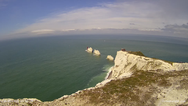 A bird's-eye view of white chalk cliffs stretching into the ocean, with a small building and a lighthouse atop the highest point, under a partly cloudy sky. Several rocks are visible protruding from the water near the cliffs.