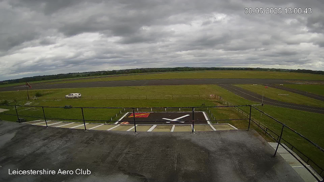 A wide view of an airport runway and taxiway under a cloudy sky. In the foreground, there is a rooftop helipad with a large white "H" and red markings, surrounded by a black safety railing. To the left, a white car is parked near a small grassy area with a windsock. In the distance, the runway extends across the landscape, with the horizon dotted by trees and low hills. The timestamp in the top right corner indicates the date as May 30, 2025, and the time as 13:00:43. The bottom left corner has text reading "Leicestershire Aero Club."