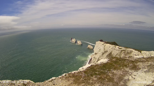 An aerial view of white chalk cliffs extending into the ocean, with a small structure on the edge of the cliff. Several large rock formations are visible in the water near the cliffs. The sky is partly cloudy with streaks of clouds stretching across the horizon.